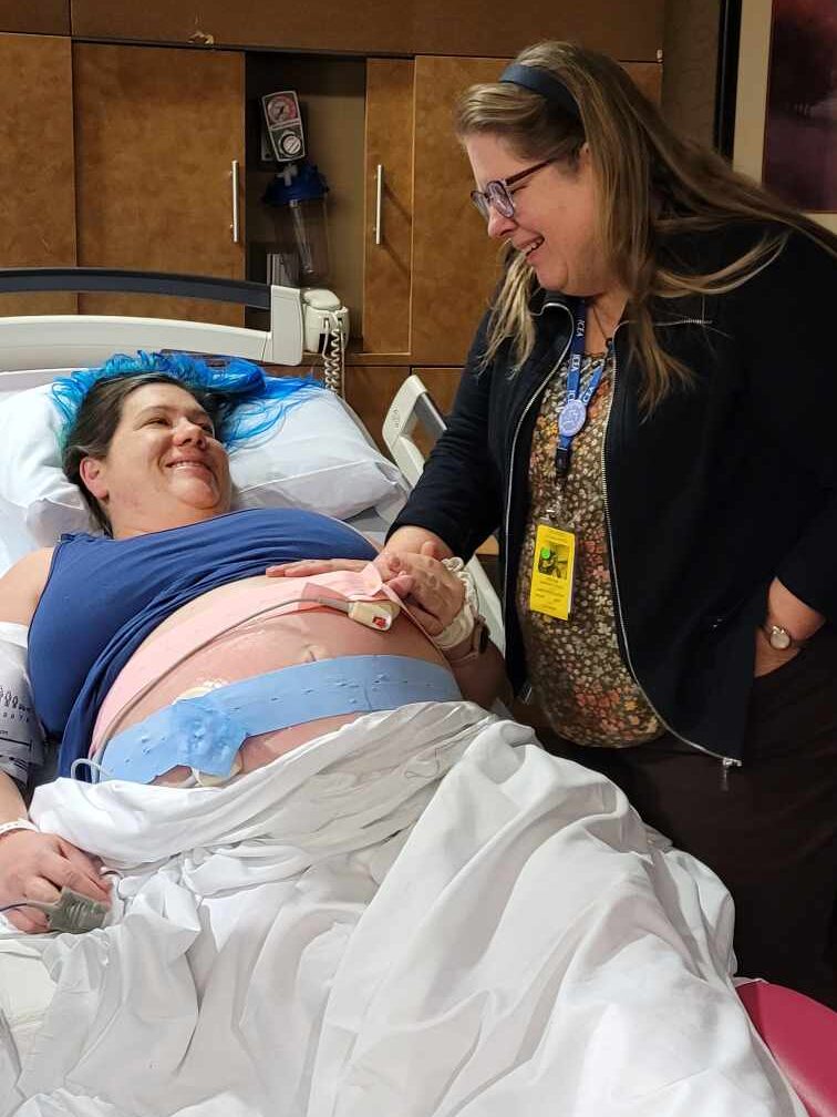 Birth Doula Stands next to a pregnant woman in a hospital bed, with a hand on the pregnant womans belly. Both women are smiling.